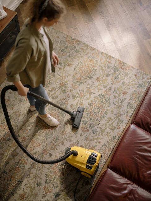 An indoor scene featuring a patterned area rug on a wooden floor, with a yellow vacuum cleaner connected to a hose, positioned near a black metal and wood TV stand. The TV stand holds a remote control