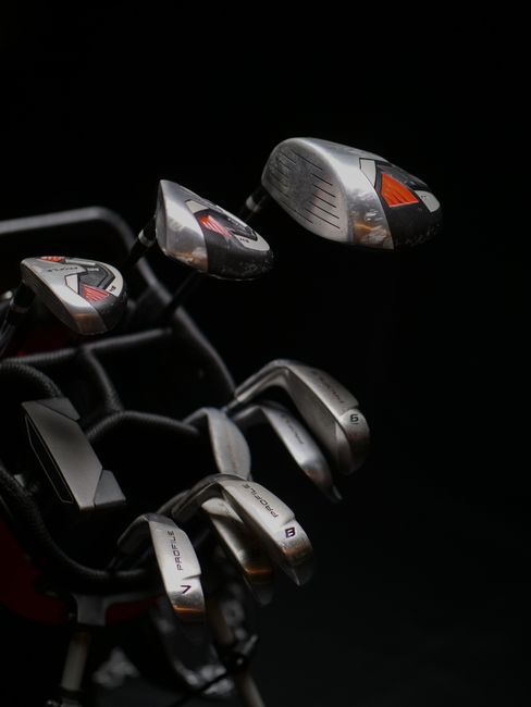 A close-up of a golf bag with several clubs, including a driver and irons, standing on a well-maintained golf course with short grass. The bag is black with blue accents and has various compartments f